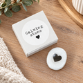 Smooth white calming worry stone with black heart detail, displayed beside its white Calming Stone gift box on a natural wooden surface.